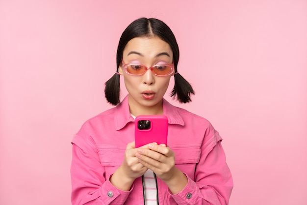 Ritratto di ragazza asiatica con occhiali da sole che utilizza lo smartphone donna che guarda il telefono cellulare che naviga nell'app in piedi su sfondo rosa