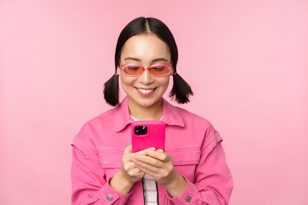 Portrait of asian girl in sunglasses using smartphone Woman looking at mobile phone browsing in app standing over pink background