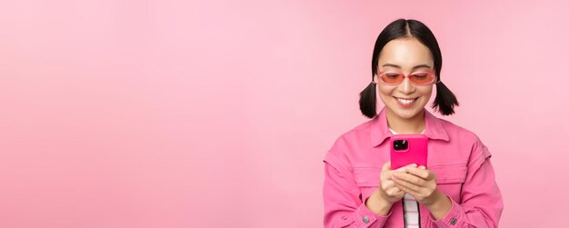 Portrait of asian girl in sunglasses using smartphone Woman looking at mobile phone browsing in app standing over pink background