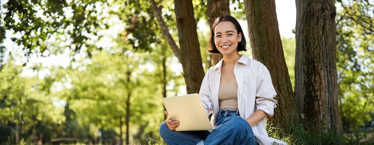 Free Photo portrait of asian girl with laptop sitting near tree in park on sunny summer day working on remote