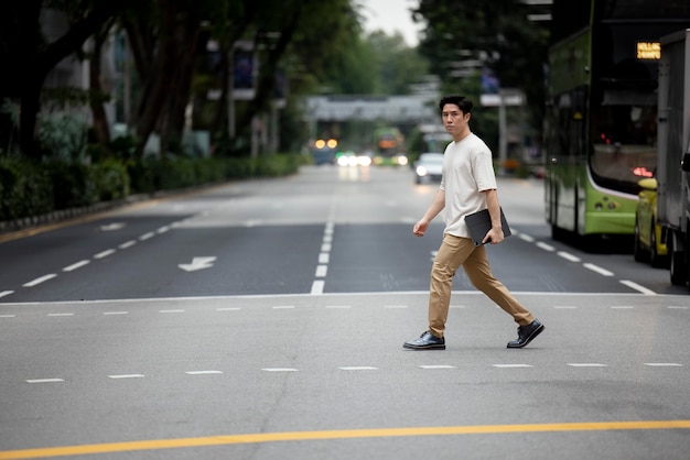 Free photo portrait of asian man crossing the street in the city while holding laptop