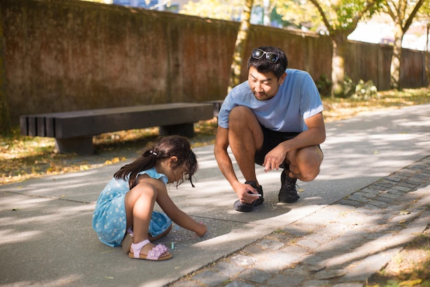 그리기를 즐기는 아시아 남자와 어린 소녀의 초상화. 행복한 아버지와 그의 딸은 공원의 포장 도로에 앉아 있고 소녀는 크레용으로 그림을 그립니다. 여가, 육아 및 건강한 성장 개념