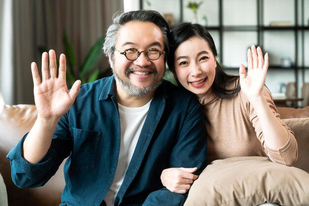リビングルームに座って笑っているアジアの成熟したカップルの肖像画妻の女性の手は後ろから夫の腕を保持し、幸せと陽気な安全なamd保険家族の概念でカメラを見てください