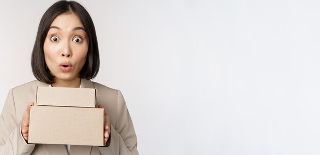 Портрет азиатской продавщицы, выглядящей удивленной, держащей коробки с доставкой товаров, стоящей в костюме на белом фоне