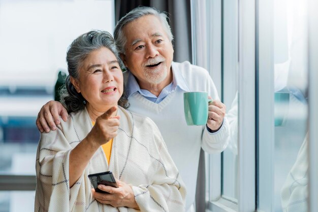 Portrait of Asian senior retired couple smiling and looking out of window apartment while hold wife shoulder Cheerful asian senior couple retirement life wellness Senior healthy lifestyle concept
