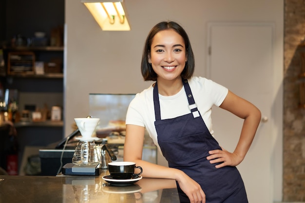 Ritratto della ragazza sorridente asiatica di barista che serve caffè che sta vicino al bancone del caffè nella preparazione del grembiule