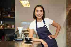 Foto gratuita ritratto della ragazza sorridente asiatica di barista che serve caffè che sta vicino al bancone del caffè nella preparazione del grembiule