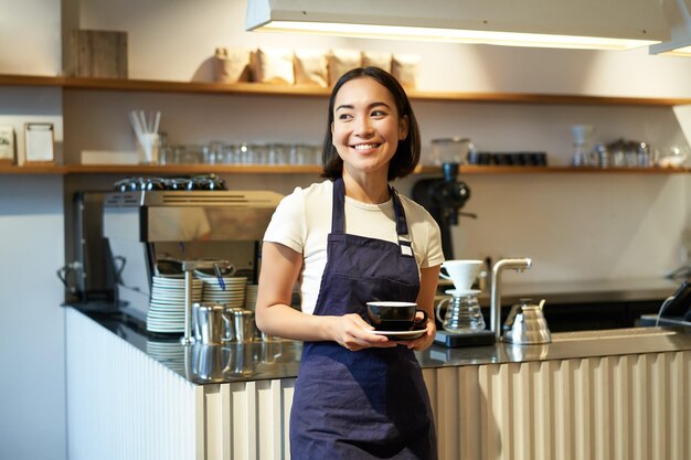 アジアの笑顔の女性バリスタ カフェ ウェイトレスの肖像画のクライアントを取るクライアントにコーヒーのカップを保持しています。