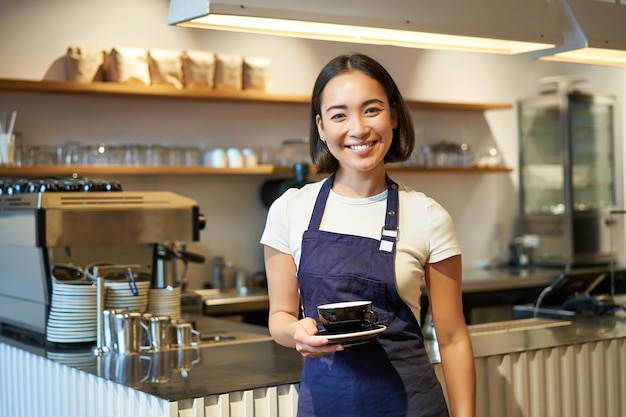 アジアの笑顔の女性バリスタ カフェ エプロンを身に着けているとクライアントを取るコーヒーのカップを保持の肖像画