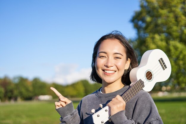 Portrait of asian smiling girl holding ukulele over shoulder pointing finger at copy space banner or