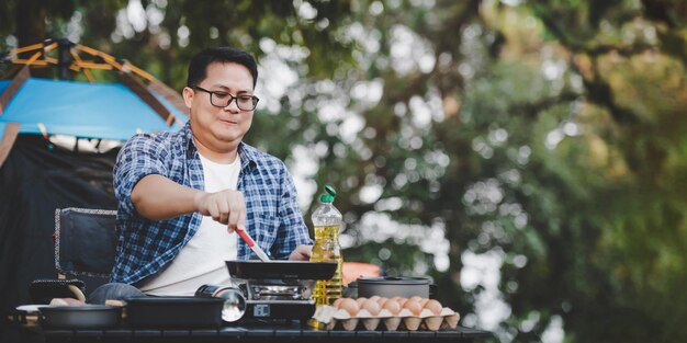 Portrait of Asian traveler man glasses pork steak frying BBQ in roasting skillet pan or pot at a campsite Outdoor cooking traveling camping lifestyle concept