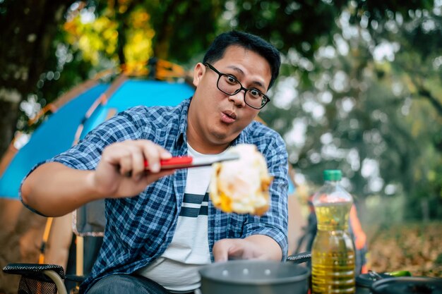 Portrait of Asian traveler man glasses pork steak frying BBQ in roasting skillet pan or pot at a campsite Outdoor cooking traveling camping lifestyle concept