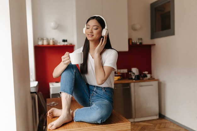 Portrait of Asian woman in denim pants and white top relaxing in headphones with cup of coffee in kitchen