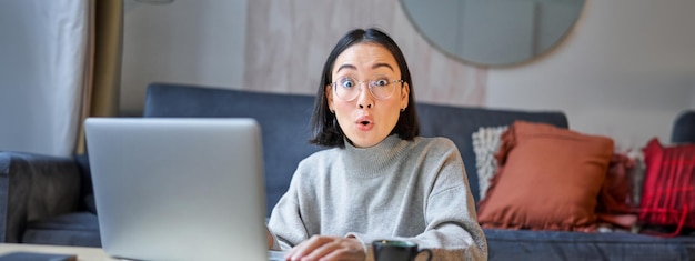 Free photo portrait of asian woman in glasses sitting with laptop and looking surprised amazed by promotion on