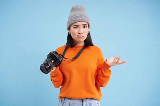 Portrait of asian woman in hat holding digital camera with confused face unprofessional photographer