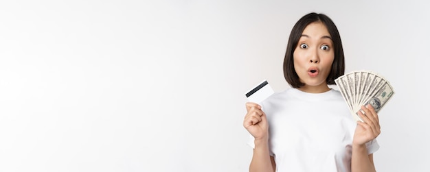 Portrait of asian woman holding money dollars and credit card looking impressed and amazed standing