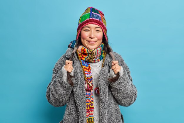 Portrait of Asian woman holds two pigtails smiles pleasantly dressed in outerwear knitted hat and scarf around neck feels happy isolated over blue wall