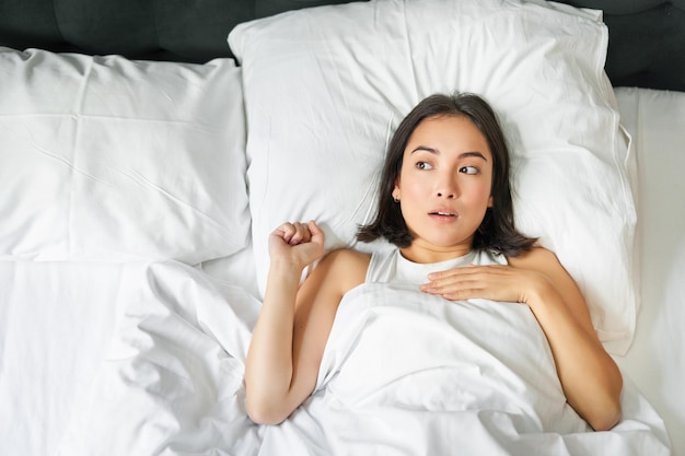 Free photo portrait of asian woman lying in bed with shocked face looking startled and upset gasping from smth