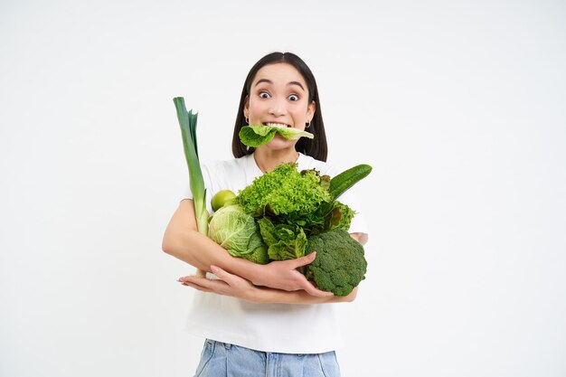 Портрет азиатки, жующей салат с зелеными органическими овощами на диетическом белом фоне