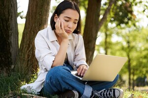 Portrait of asian woman sitting on grass near tree using laptop working doing homework remotely on s