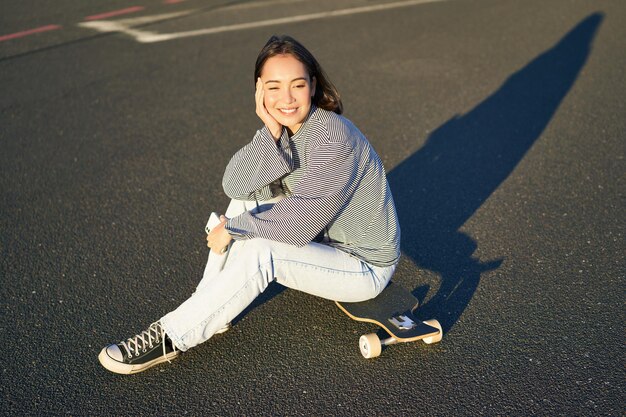 Портрет азиатской женщины, сидящей на скейтборде и катающейся на своем круизном лонгборде с помощью приложения для смартфона