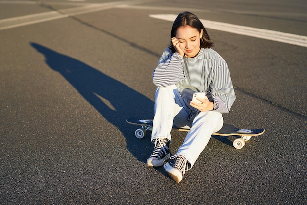 Портрет азиатской женщины, сидящей на скейтборде и катающейся на своем круизном лонгборде с помощью приложения для смартфона