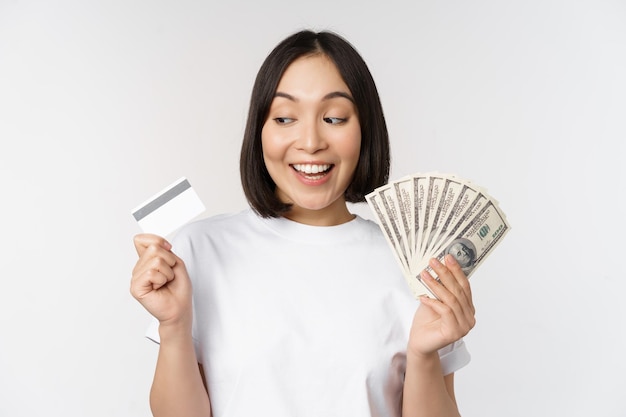 Portrait of asian woman smiling holding credit card and money cash dollars standing in tshirt over white background