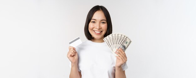 白い背景の上にtシャツに立っているクレジットカードとお金の現金ドルを持って笑っているアジアの女性の肖像画