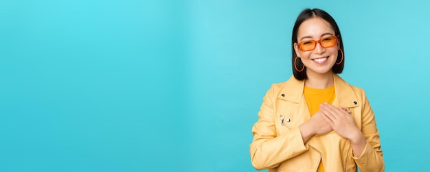 Portrait of asian woman smiling holding hands on heart and looking with tenderness care at camera thankful emotion standing over blue background