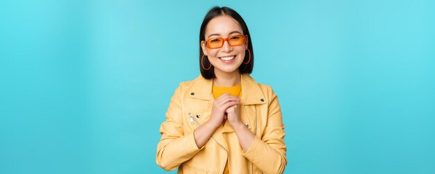 Portrait of asian woman in sunglasses looking hopeful flattered smiling happy standing over blue background
