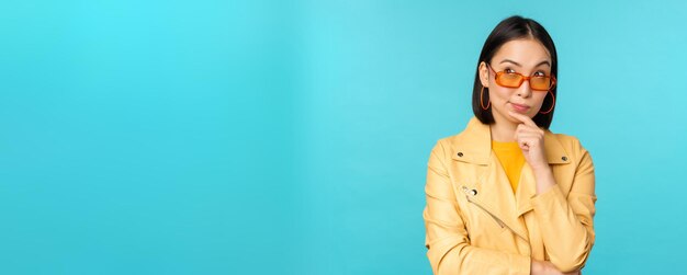 Portrait of asian woman thinking looking thoughtful searching ideas or solution wearing sunglasses standing over blue background