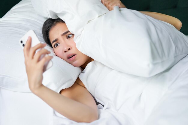 Free photo portrait of asian woman waking up in bed looking shocked at mobile phone realise she overslept