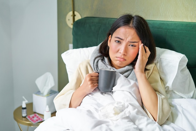 ベッドの飲み物に横たわっている自宅で病欠に滞在しているコルを捕まえる頭痛を持つアジアの女性の肖像画