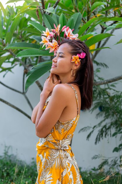 Portrait of asian woman in yellow summer dress stands with plumeria thai flower in hair and round earings Female with light make up outside on background of wall and green bushes