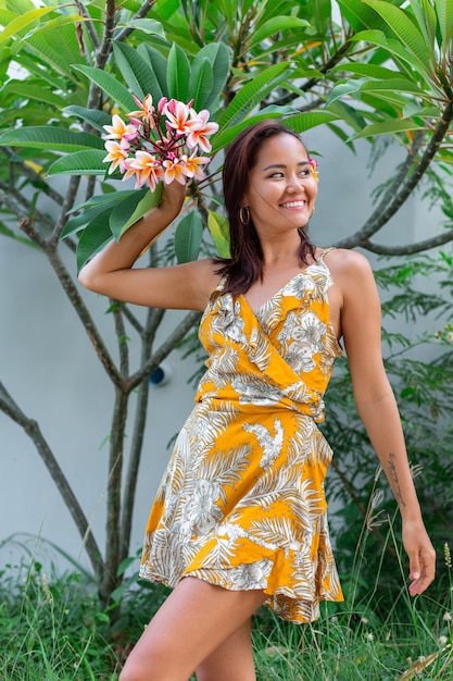 Free photo portrait of asian woman in yellow summer dress stands with plumeria thai flower in hair and round earrings