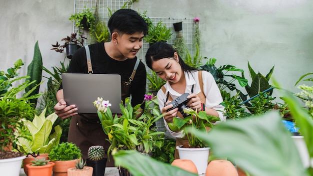 エプロンを身に着けている肖像画アジアの若い庭師のカップルは、温室内の観葉植物の世話をしながら写真を撮るためにラップトップとカメラを使用しています