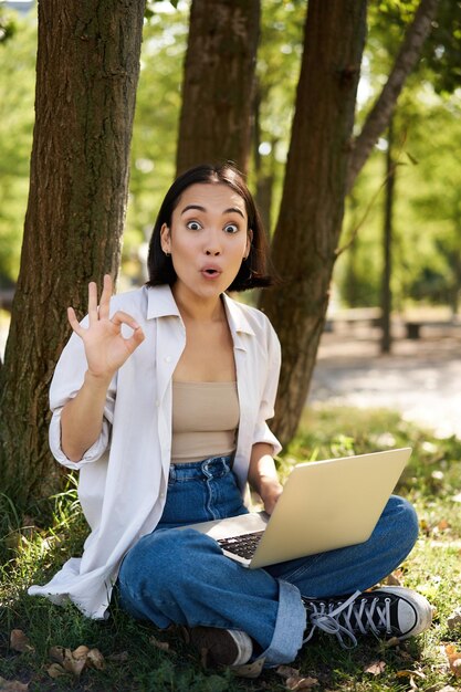 ラップトップで木のそばに座って公園で働く宿題をしているアジアの若い女性学生の肖像画