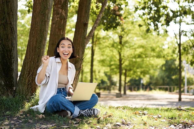 Portrait of asian young woman student doing homework working in park sitting beside tree with laptop