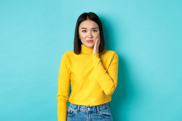 Portrait of asian young woman touching cheek and frowning, looking sad, being slapped in face, feeling painful toothache, standing over blue background