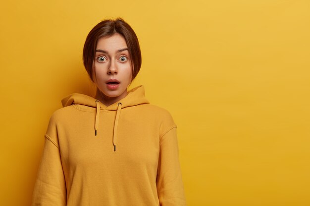 Portrait of astonished beautiful female hipster holds breath from excitement and surprisement, stares with bugged eyes, wears yellow casual hoodie