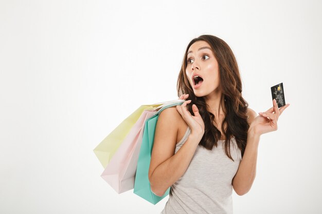 Portrait of astonished woman with lots purchases in hand doing shopping using credit card, isolated over white