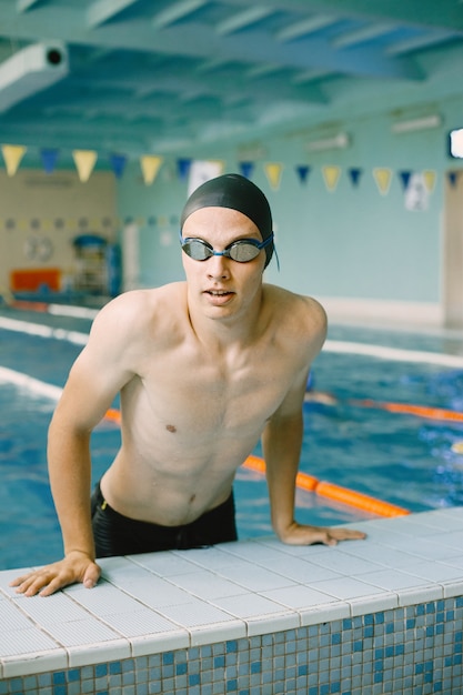 Портрет пловца спортсмена в кепке сбоку в бассейне. Пловец в закрытом бассейне выходит из воды.