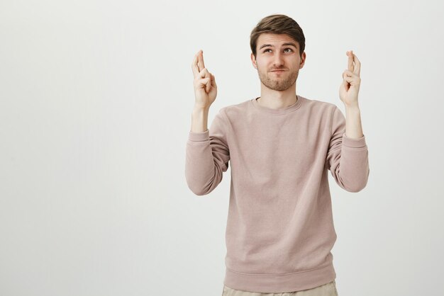 Portrait of athletic unshaved guy standing with raised hands and crossed fingers looking up with determined face hoping to avoid something over gray wall