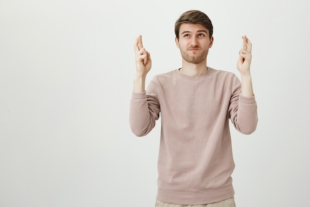 Portrait of athletic unshaved guy standing with raised hands and crossed fingers looking up with determined face hoping to avoid something over gray wall