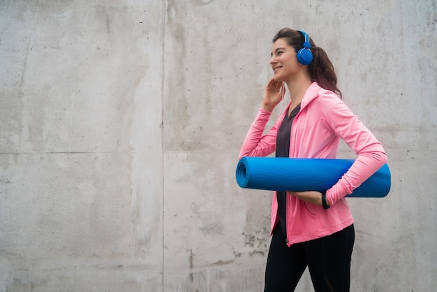 音楽を聴きながらトレーニングマットを保持している運動女性の肖像画。スポーツとライフスタイルのコンセプト。