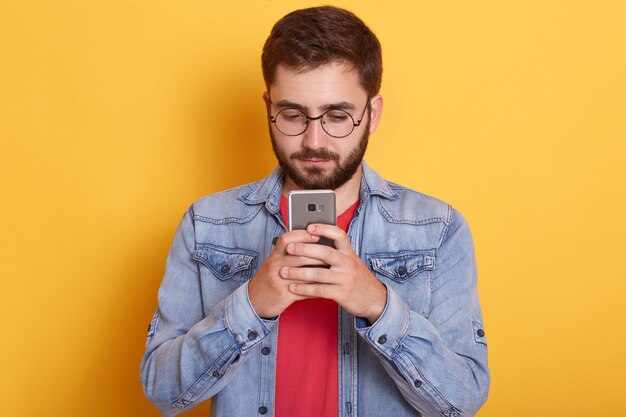 Portrait of attentive serious handsome man holding his smartphone in both hands, looking at its screen, being interested in device