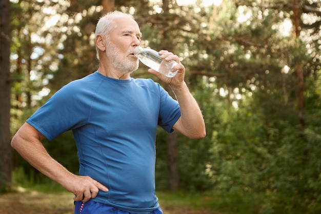 Portrait of attractive active male pensioner with bald head and stubble refreshing himself after jogging outdoors, standing against pine forest, holding bottle of drinking water