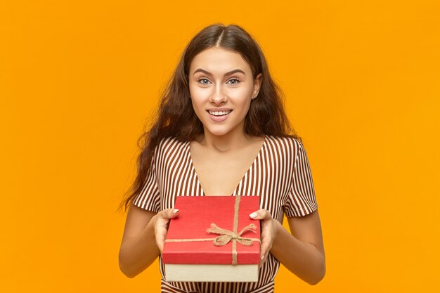 Portrait of attractive adorable young european female in striped dress holding red box