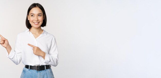 Портрет привлекательной взрослой азиатской женщины, указывающей налево с довольной улыбкой, показывающей баннер или логотип в стороне, стоящей на фоне белой студии