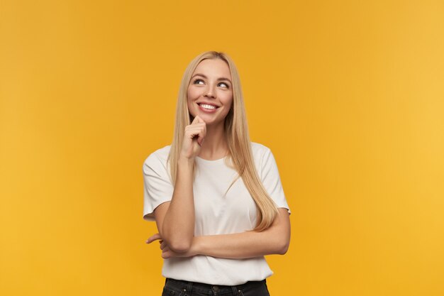 Portrait of attractive, adult girl with blond long hair. wearing white t-shirt and black jeans. people and emotion concept. watching up at copy space, isolated over orange background